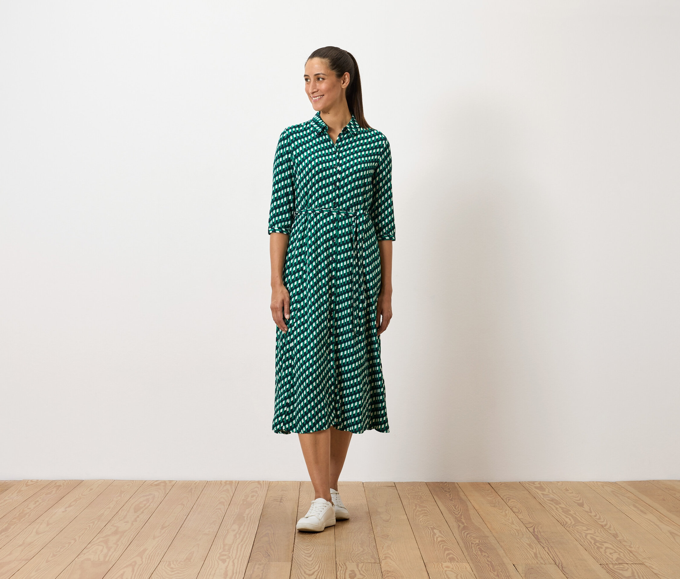 Egy nő áll, zöld mintás ruhát és fehér tornacipőt visel.