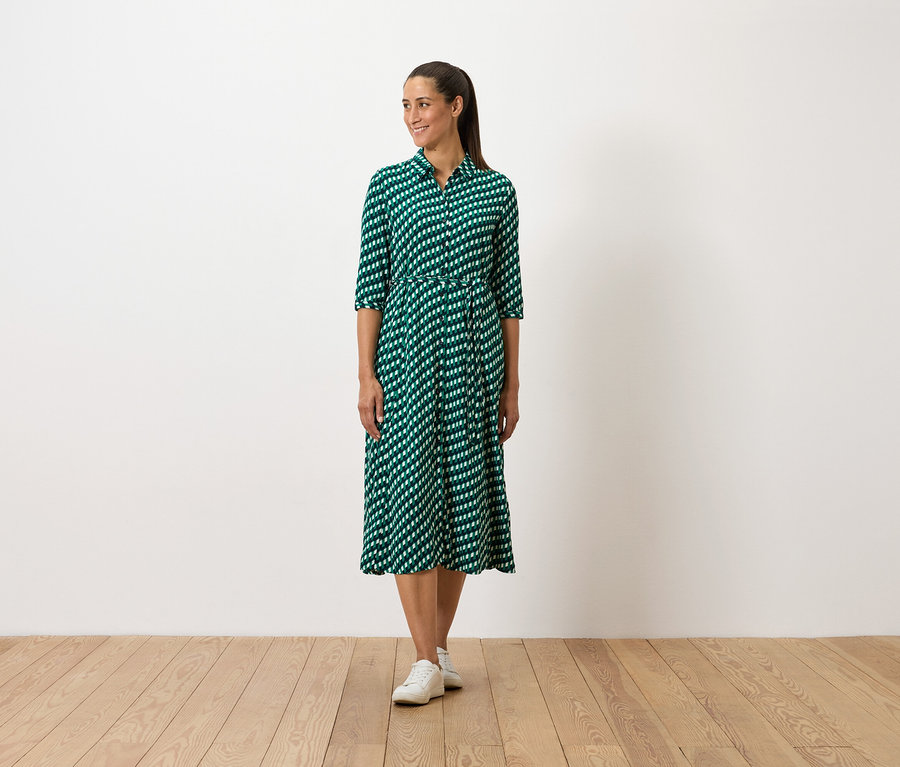 Egy nő áll, zöld mintás ruhát és fehér tornacipőt visel.