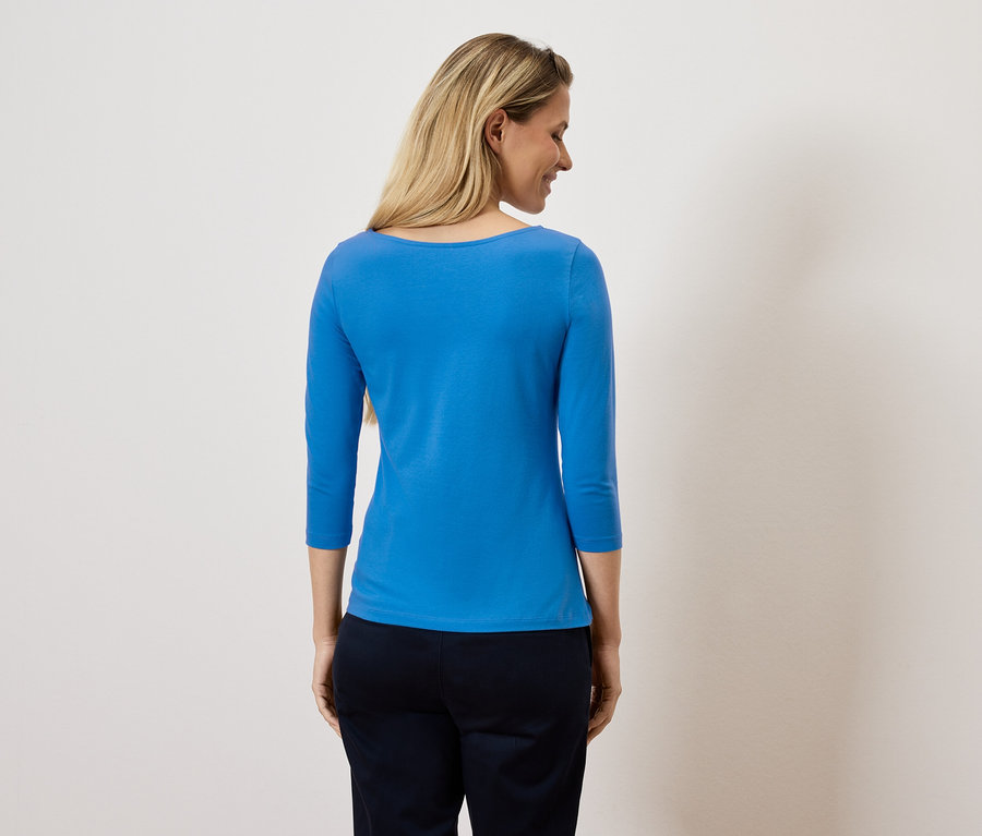 Egy nő a válla fölött néz, kék háromnegyedes ujjú felsőt és sötét nadrágot visel.