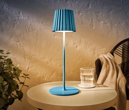 Egy kék LED-es akkumulátoros kültéri asztali lámpa áll egy asztalon egy pohár víz mellett.