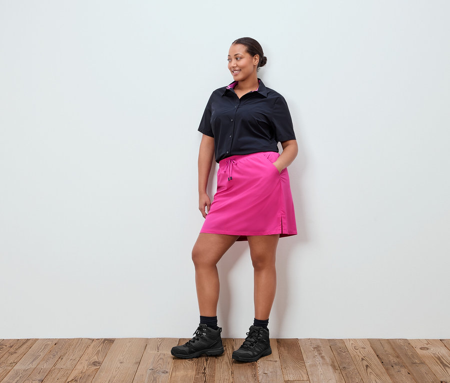 Egy nő áll egy fehér fal előtt, fekete inget, rózsaszín szoknyát és fekete cipőt visel.