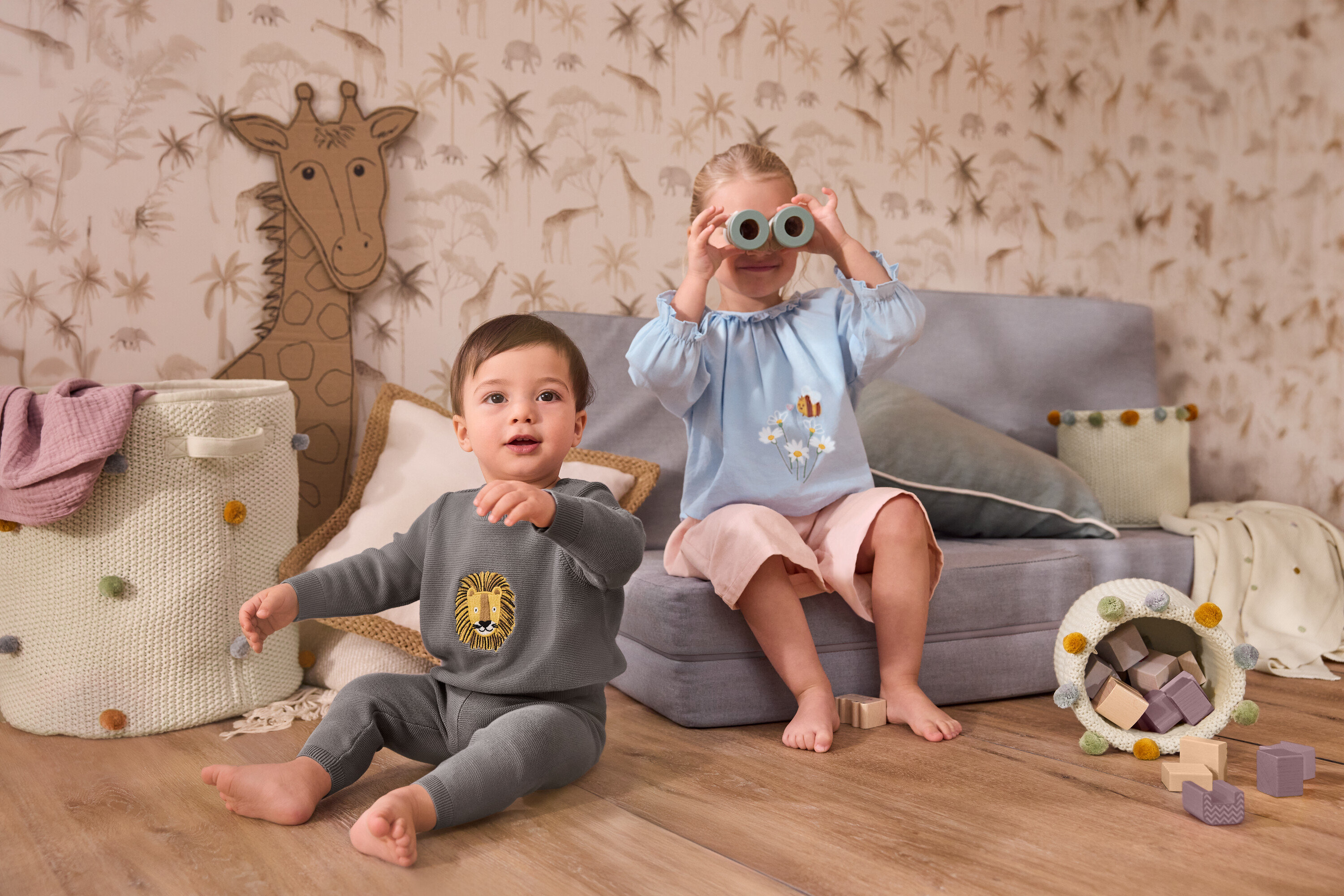 Egy kisgyermek ül egy nagy gyerekszobai tárolókosár mellett. Egy lány egy szürke kanapén ül, és egy kötött babatakarót tart az arca elé. Gyerek chambray blúzt és gyerek kordbársony culotte-ot visel.