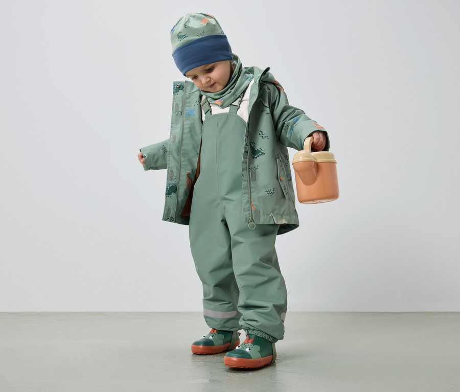 Egy dinoszaurusz mintás, zöld esőruhát viselő gyermek narancssárga vödröt tart.