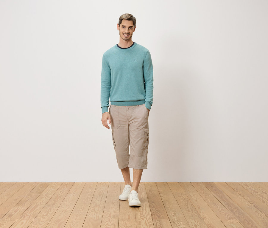 Egy férfi áll zsebre tett kézzel egy fehér fal előtt. Világoskék pulóvert, bézs színű háromnegyedes cargo nadrágot és fehér cipőt visel.