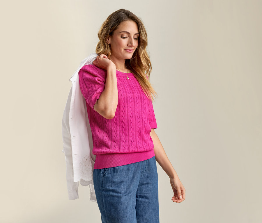 Nő a vállán farmerdzsekivel, rózsaszín, finomkötésű, fonott mintás pulóverben és farmernadrágban.