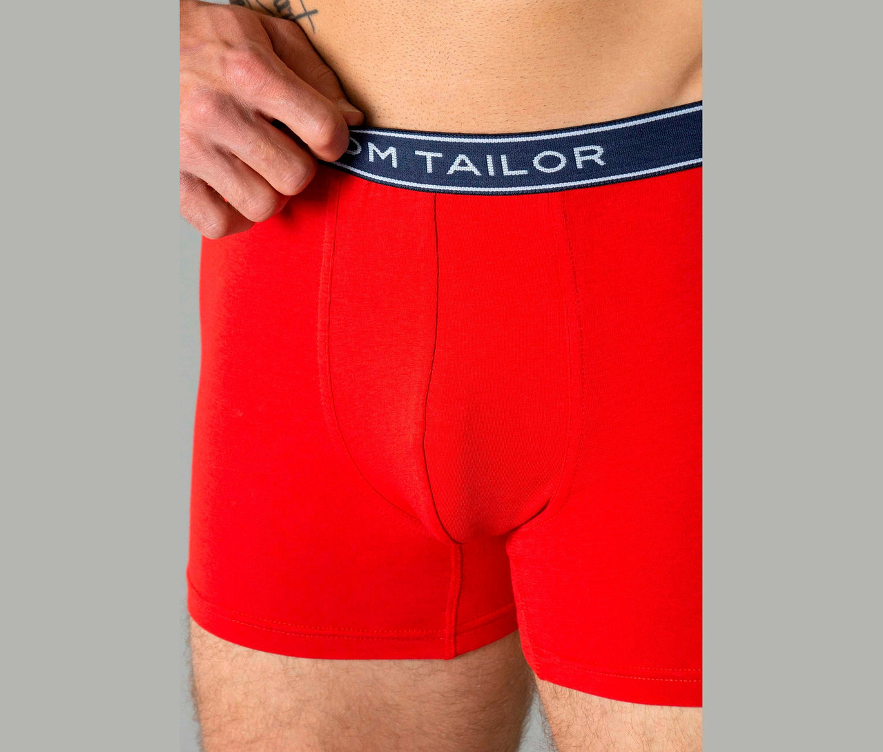 Egy férfi piros boxeralsót visel, sötétkék derékpánttal, rajta a „TOM TAILOR” felirat.