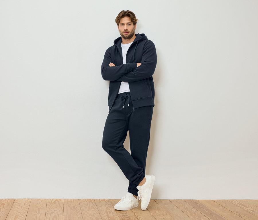 Egy férfi keresztbe font karokkal, tengerészkék kapucnis cipzáras pulóvert, tengerészkék melegítőnadrágot és fehér tornacipőt visel, egy fehér fal előtt állva.