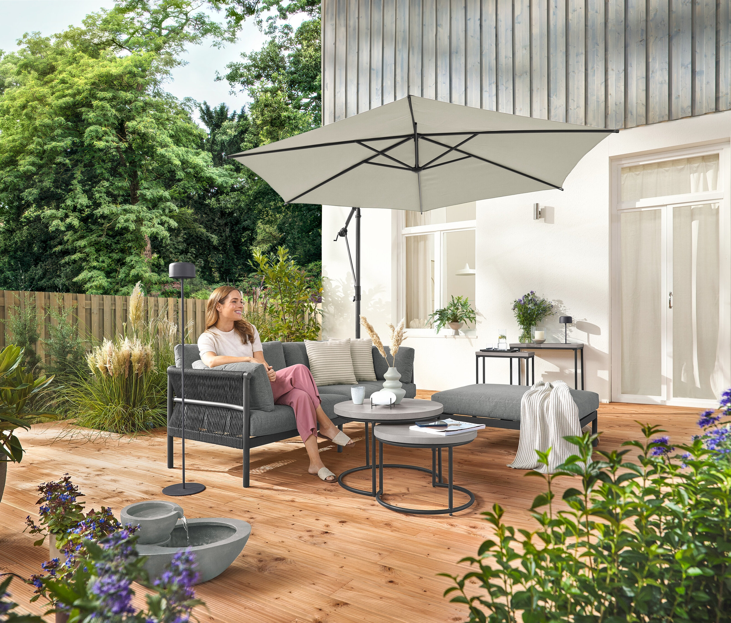 Egy nő ül egy kényelmes kerti kanapén, különálló lábtartóval. Mellette egy szürke napelemes akkumulátoros állólámpa áll. Elöl egy napelemes kerti szökőkút.