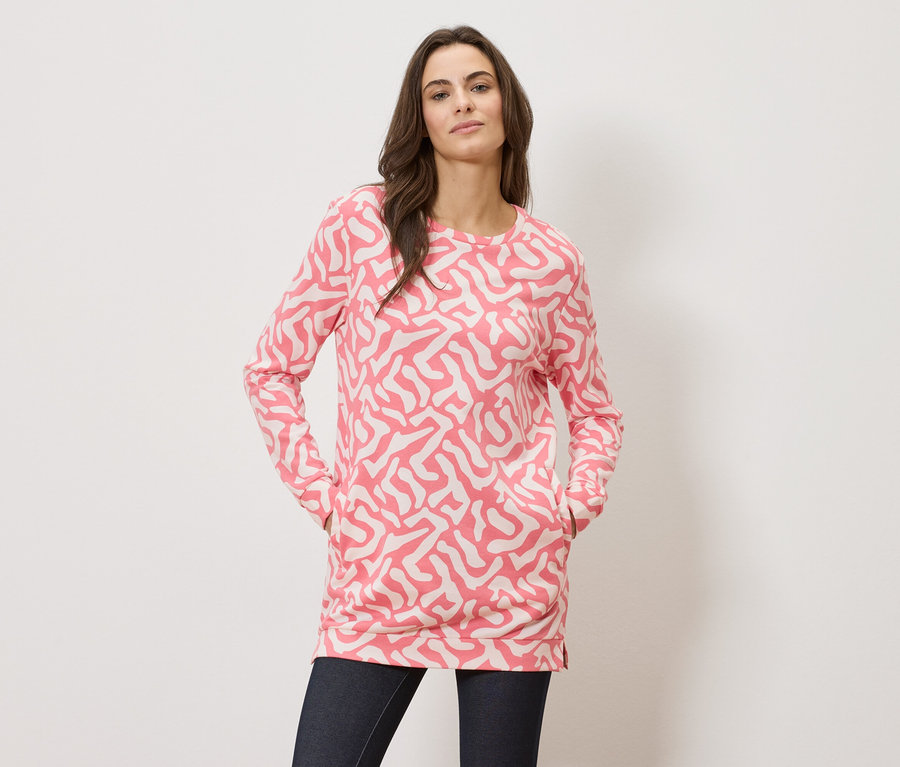 Egy nő a zsebében tartja a kezét, rózsaszín mintás, hosszú ujjú felsőt és sötét farmert visel.