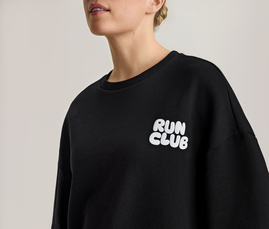 Nő fekete szabadidőfelsőt visel, fehér "RUN CLUB" felirattal.
