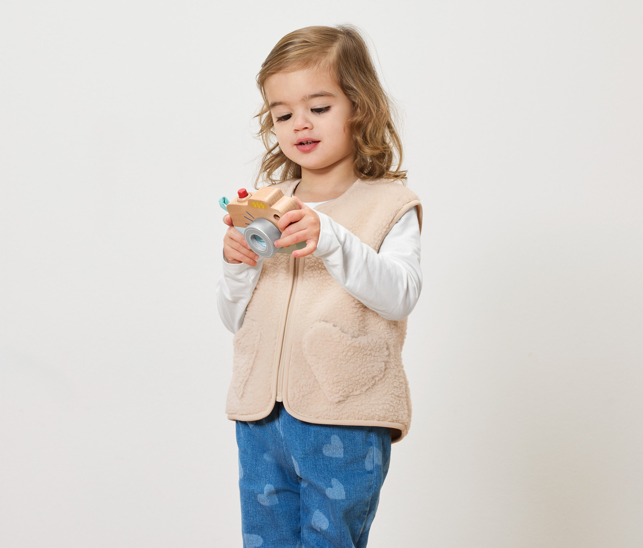 Egy göndör hajú kislány játék fényképezőgépet tart a kezében világos háttér előtt.