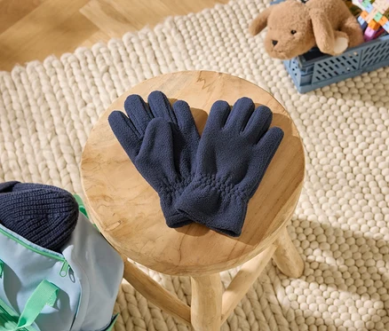 Sötétkék polárkesztyűk egy fa sámlin. Mellette egy világoskék táska sötétkék sapkával. A háttérben egy plüssmackó egy kék ládában, egy szőnyegen.