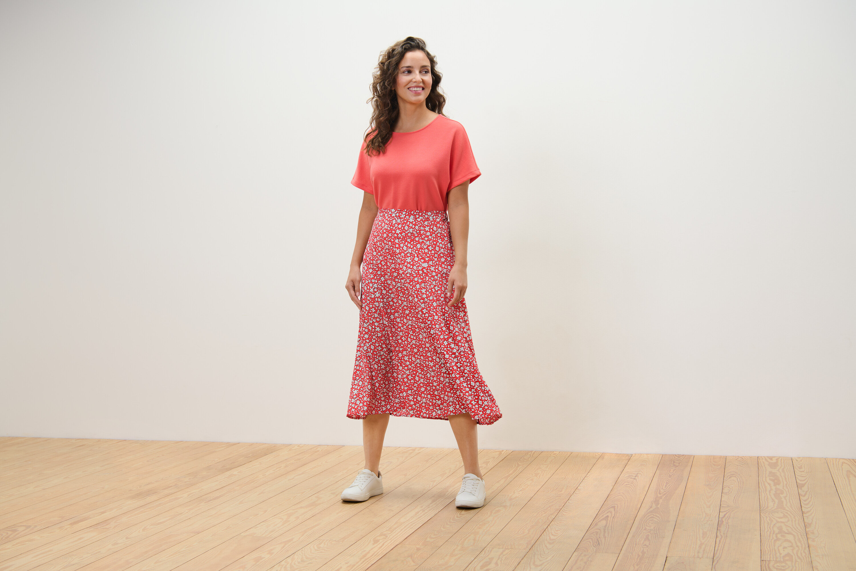 Nő áll a fapadlón, korall színű, mintás midiszoknyát visel.