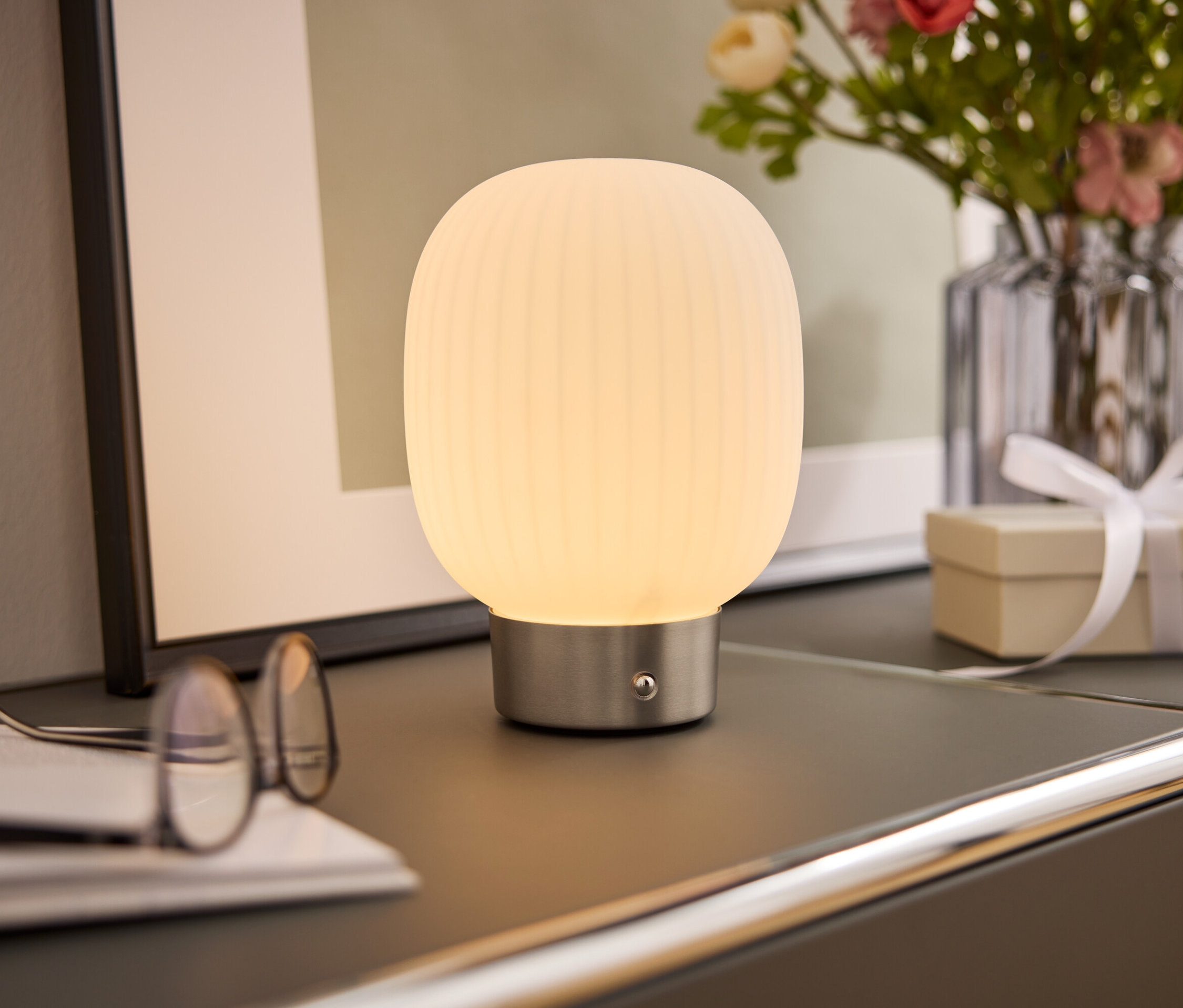 Éjjeli lámpa fehér lámpaernyővel áll egy szekrényen egy szemüveg, egy könyv, egy virágváza és egy ajándékdoboz mellett.