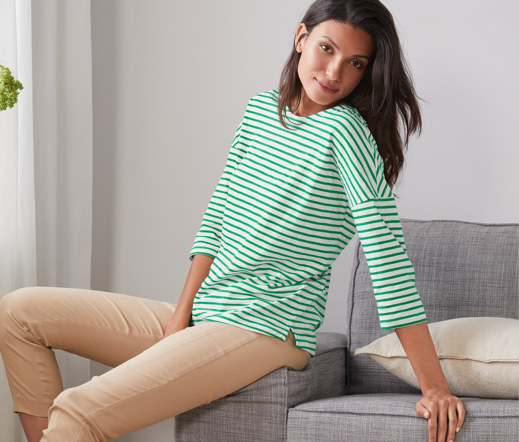 Nő ül egy szürke kanapén, és Női denevérujjú felső, csíkos, zöld/fehér felsőt visel.