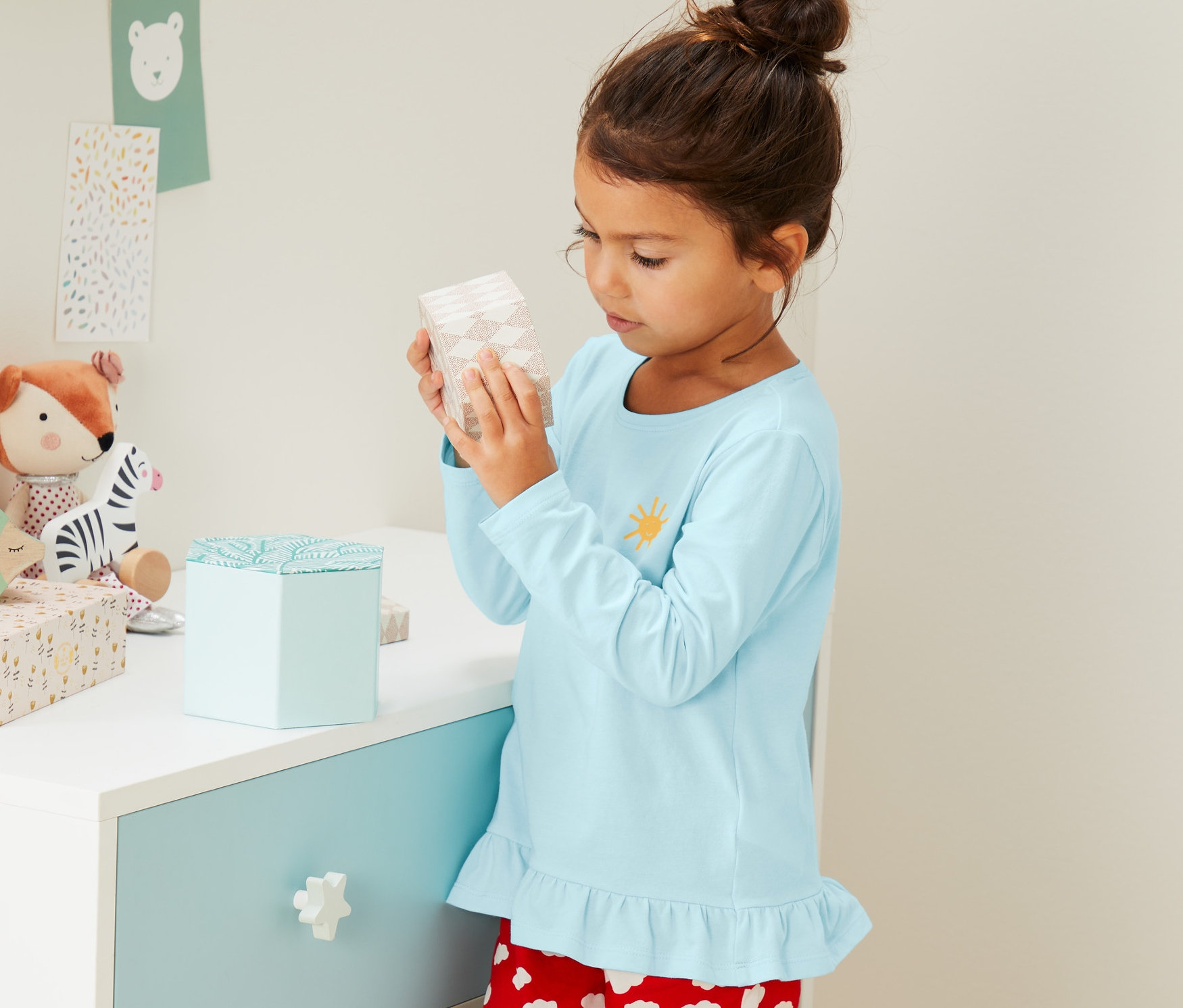 Egy lány díszdobozt tart a gyerekszobában. Világoskék felsőt és felhős, piros nadrágot visel.