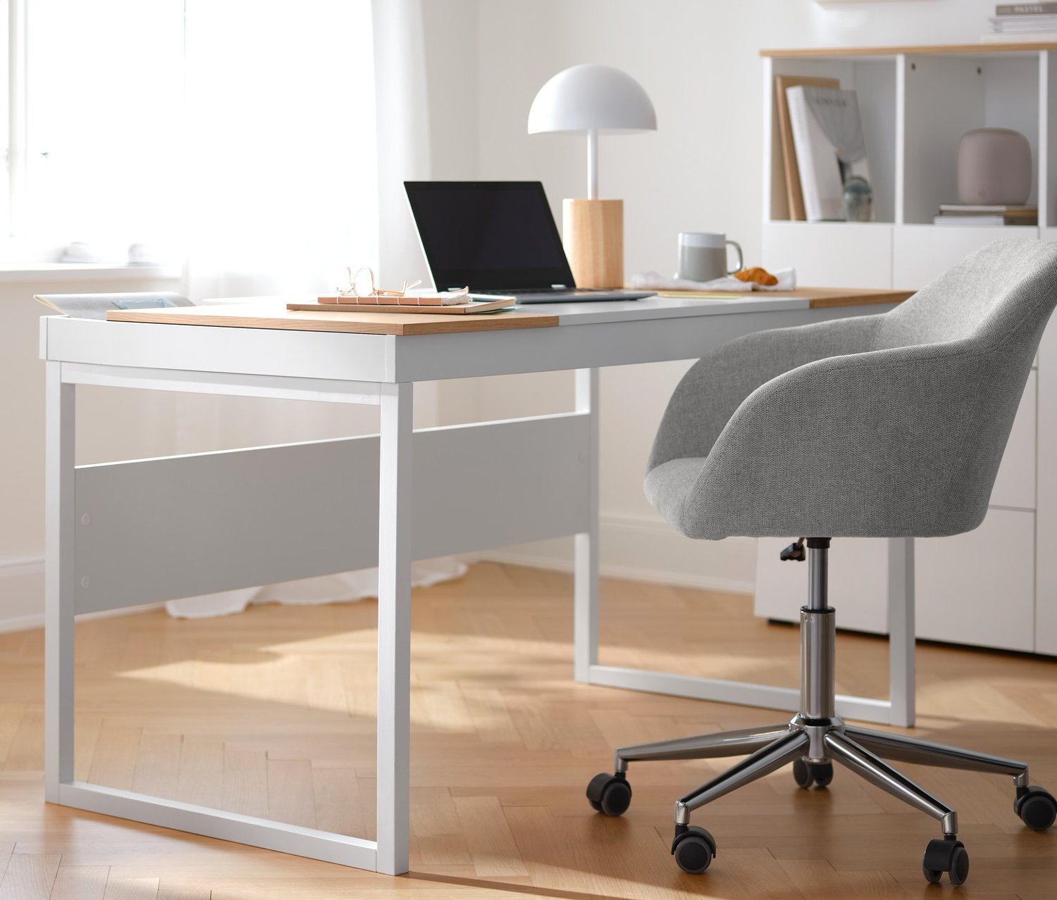 Egy bézs irodai szék áll egy fehér íróasztalnál. Balra egy görgős tároló, tálcával látható. A háttérben egy szekrény nyitott polcokkal áll.