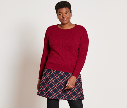 Egy nő áll, piros kötött pulóvert, kockás jacquard szoknyát, fekete, 2 darabos harisnyanadrágot és fekete bőrcsizmát visel.