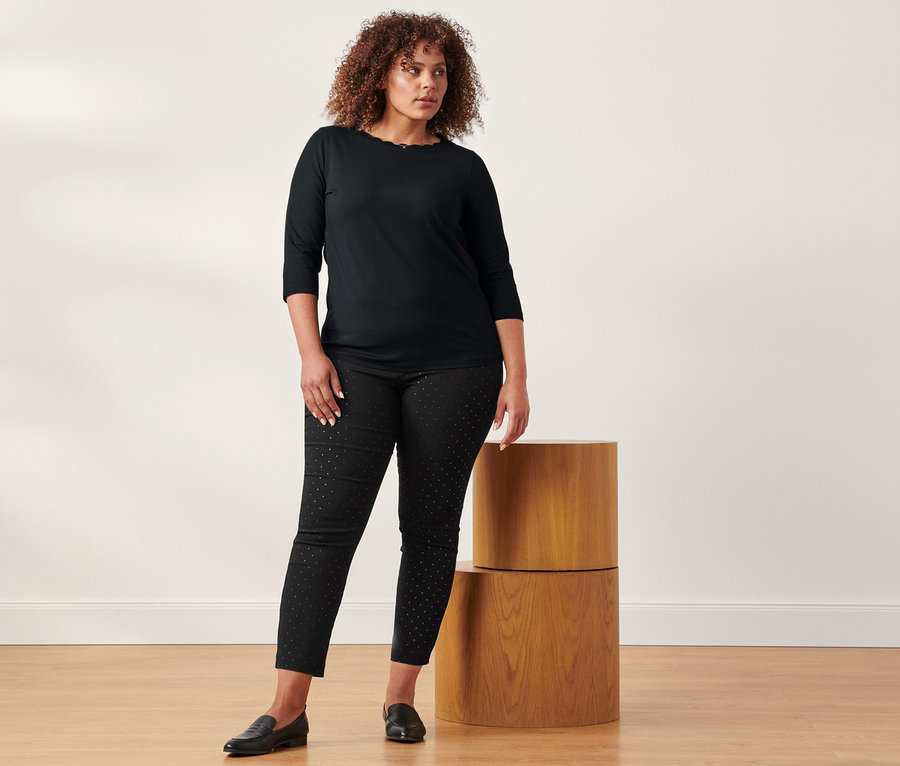 Egy nő áll, fekete, 3/4-es ujjú felsőt és pöttyös sztreccsnadrágot visel.