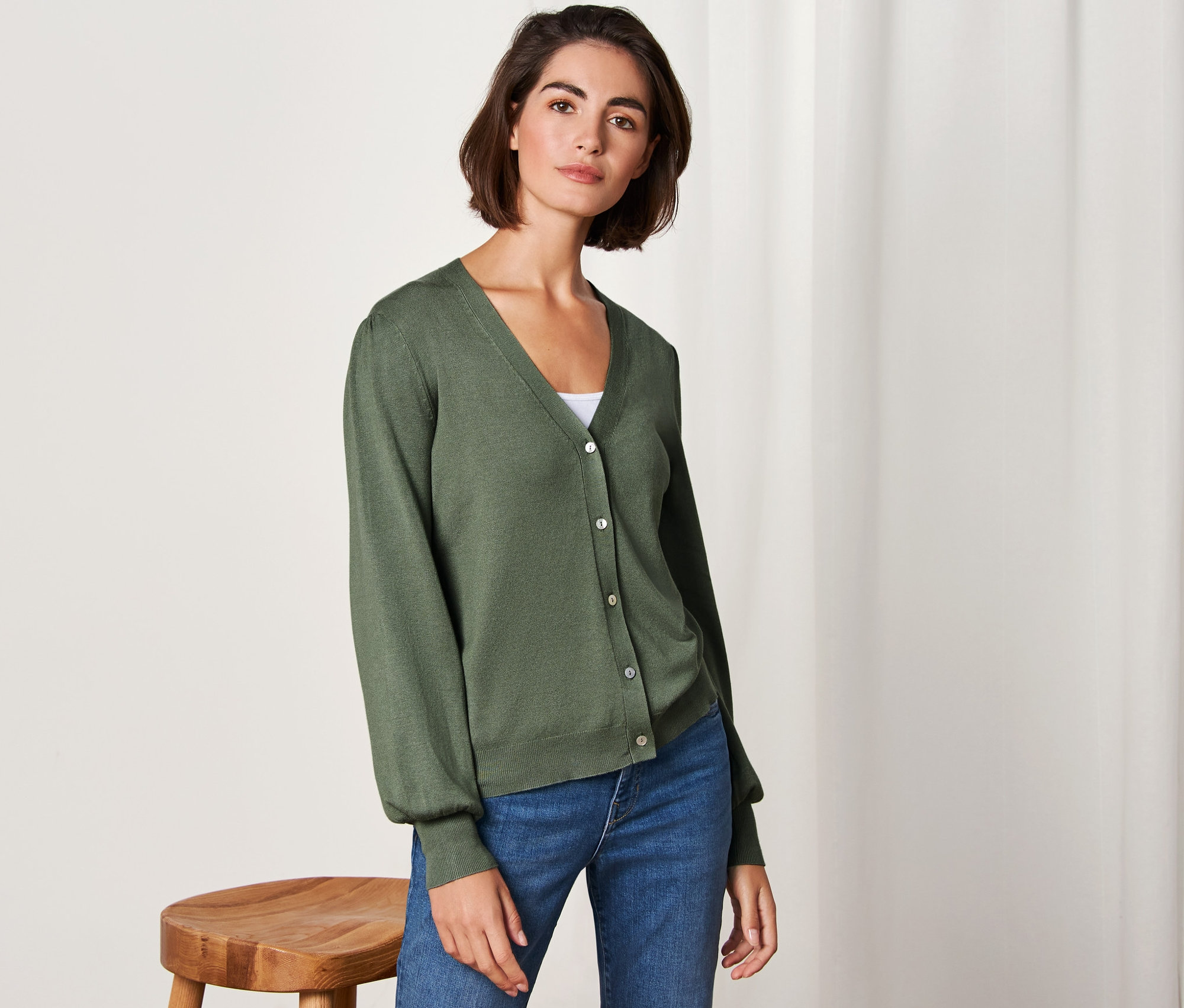 Nő pózol zöld, finomkötésű kardigánban és farmerban.