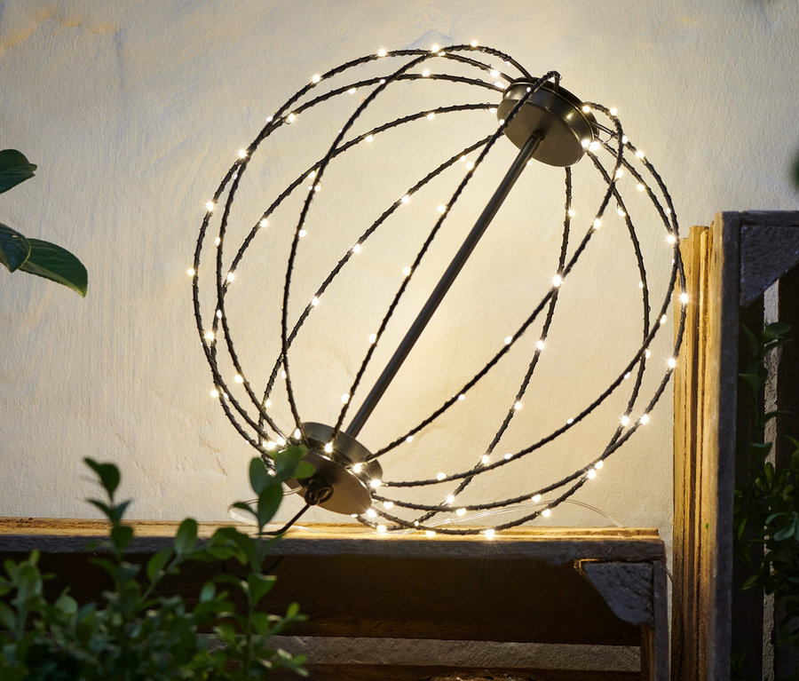 Egy fém LED gömb áll egy fa ládán növények mellett.