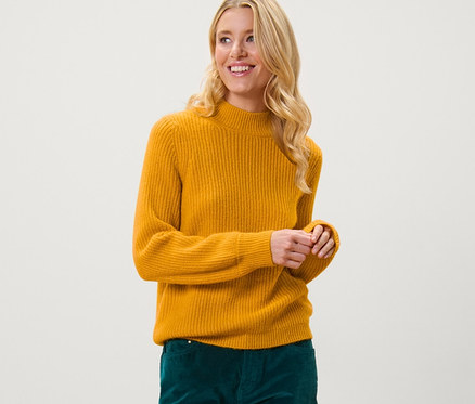 Szőke nő mustársárga, laza kötésű pulóverben és zöld nadrágban pózol.