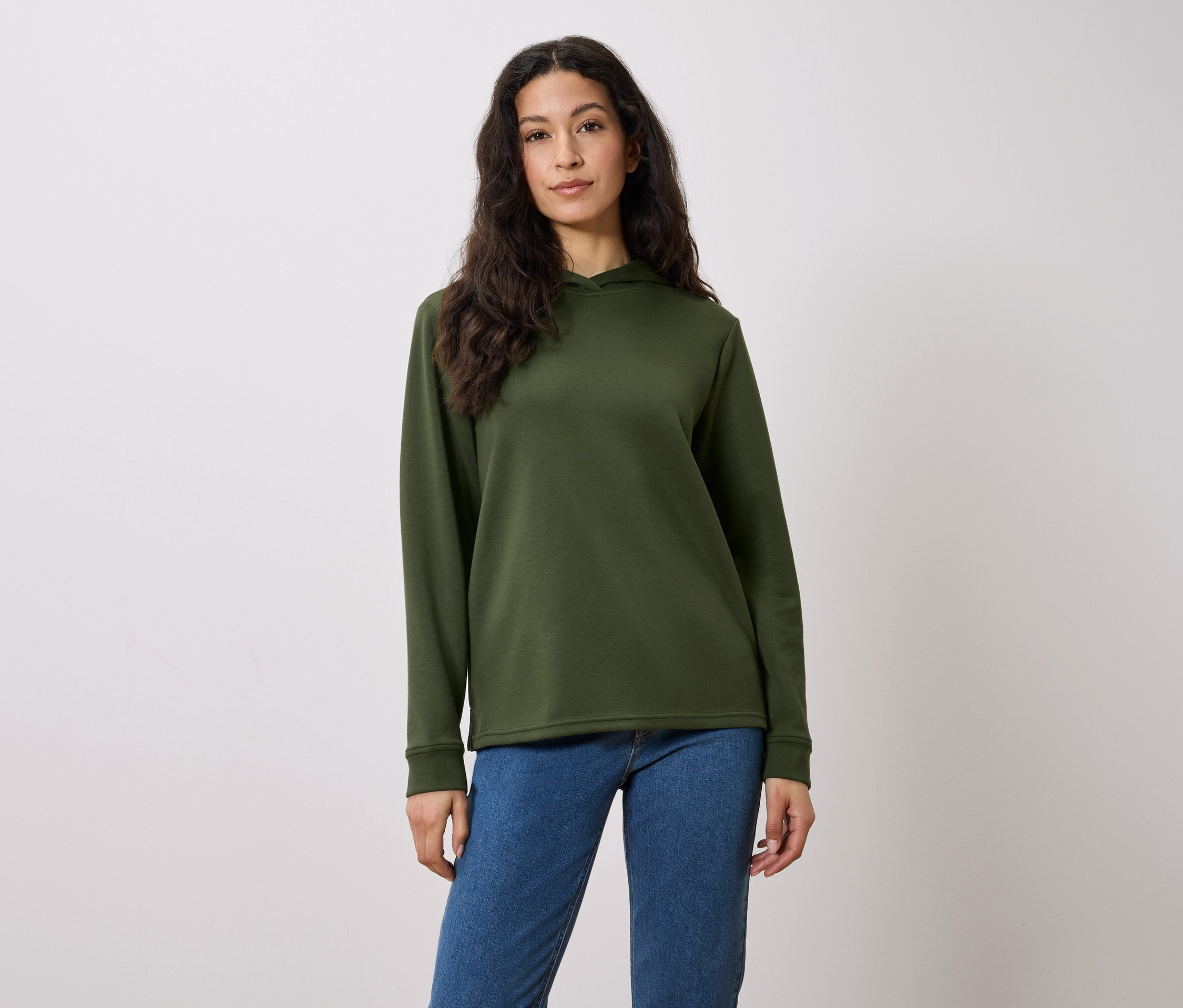 Egy hosszú, barna hajú nő olívazöld kapucnis pulóverben és kék farmerben pózol fehér háttér előtt.