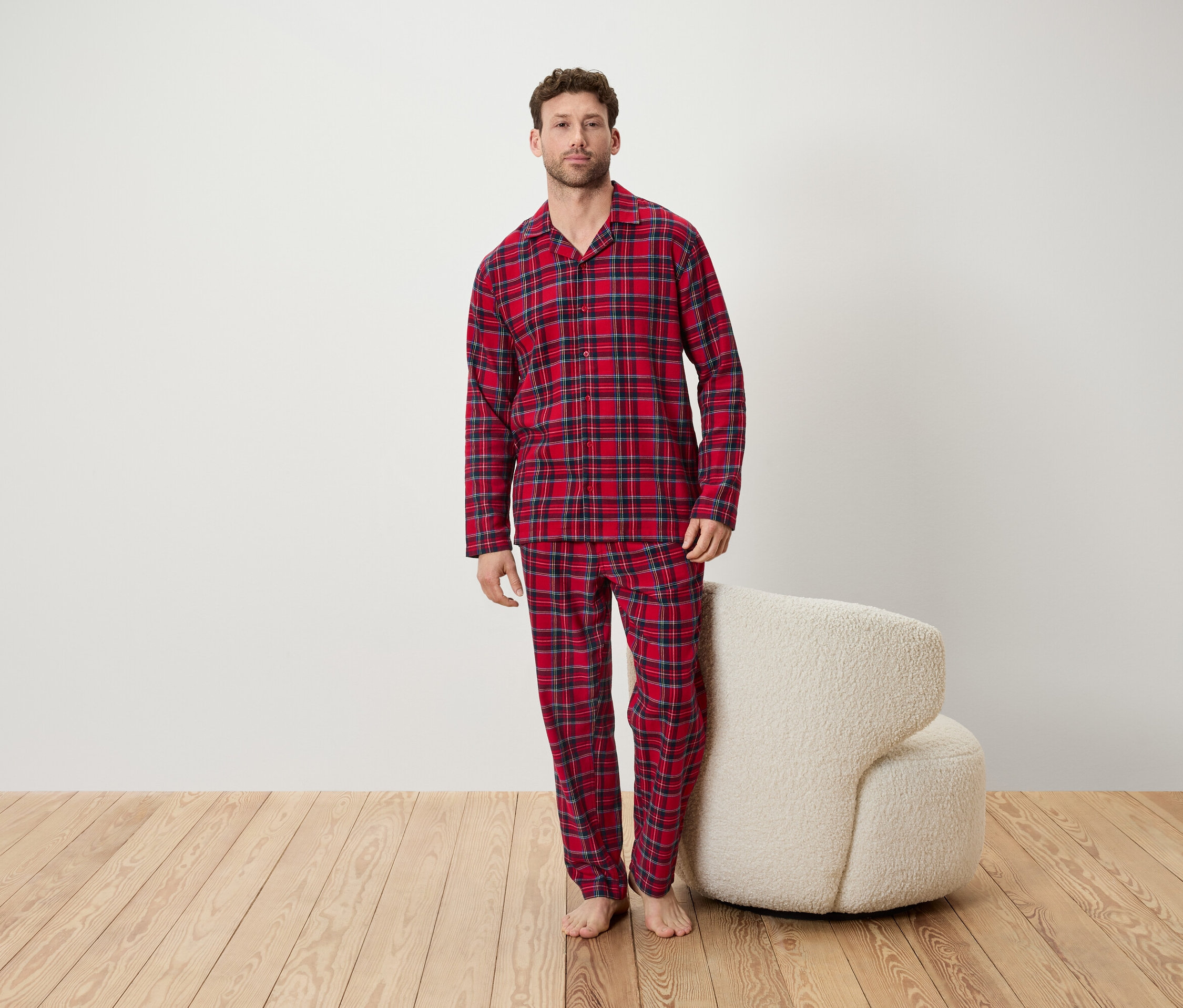 Férfi piros kockás pizsamában áll egy szobában egy fehér szék mellett.