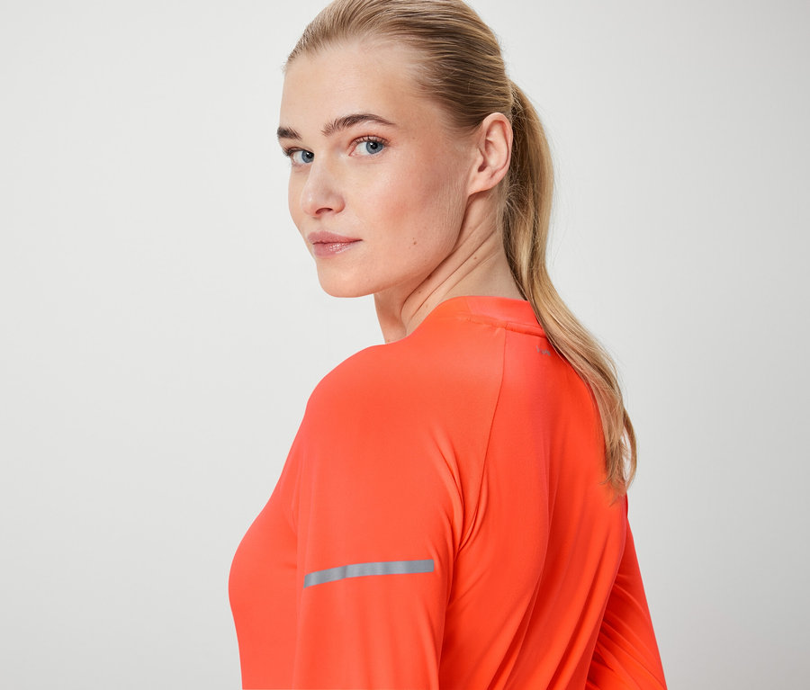 Egy szőke nő a válla fölött néz, narancssárga, hosszú ujjú felsőt visel.