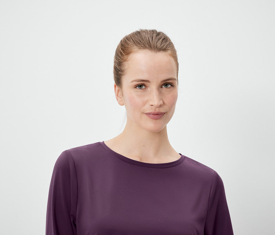 Portré egy lófarokba kötött hajú nőről, aki lila, hosszú ujjú felsőt visel, fehér háttér előtt.