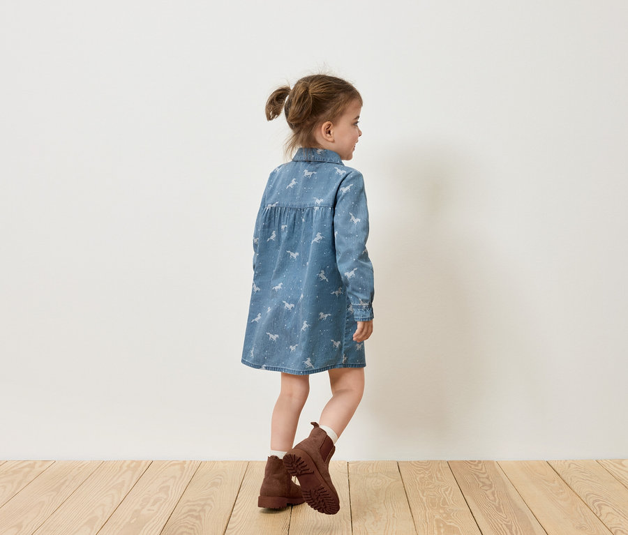 Kislány ló mintás farmerruhában és barna csizmában áll oldalt egy fehér fal előtt.