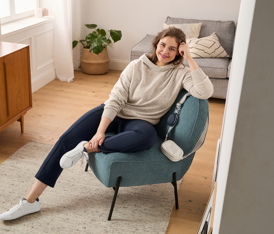 Egy nő ül egy fotelben, és finomkötésű pulóvert, bőszárú leggingst és fehér sneakert visel. A fotelre oldaltáska van akasztva.