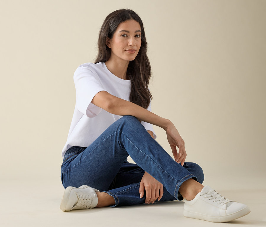 Nő ül a földön, Slim-Jeans – Fit »Emma« farmerben, középkék farmer színben, fehér pólóban és tornacipőben.
