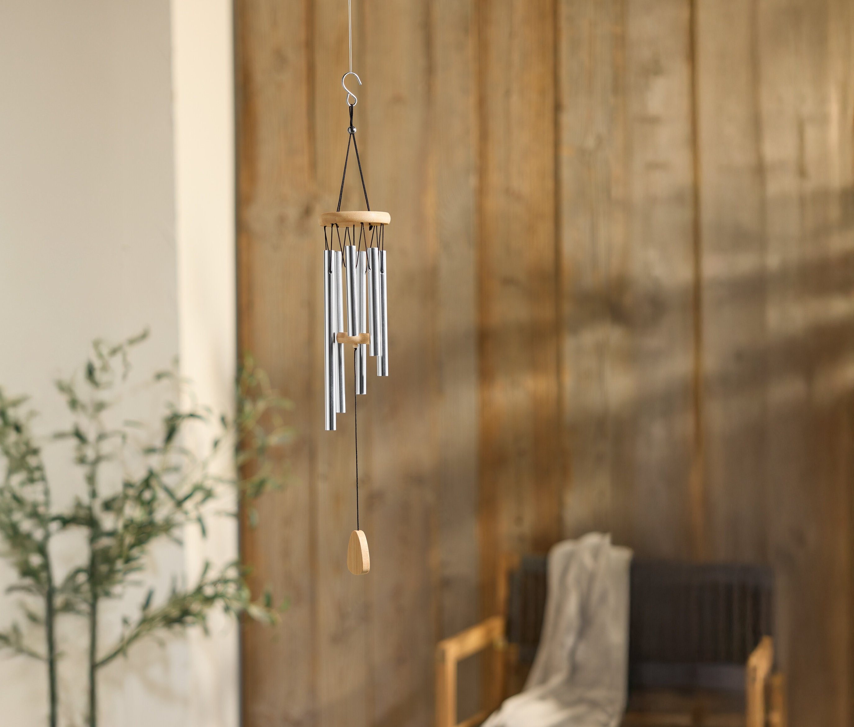 Egy ezüst színű csövekkel ellátott szélcsengő lóg egy fa fal előtt, egy növény és egy szék mellett.