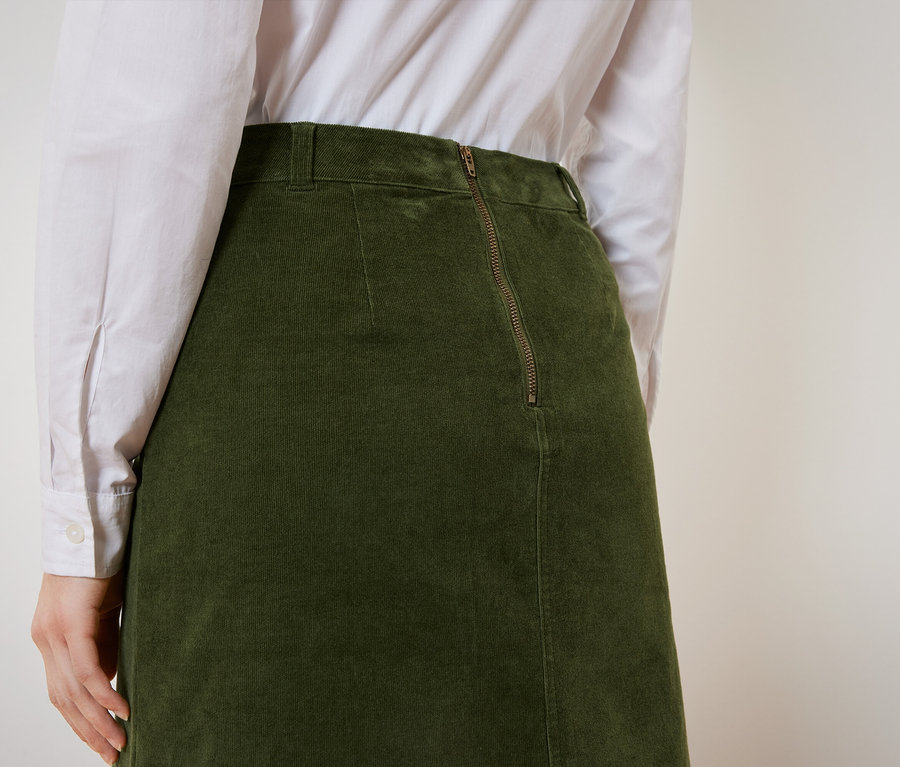 Hátulnézet egy olívazöld kordbársony szoknyát és fehér inget viselő nőről.