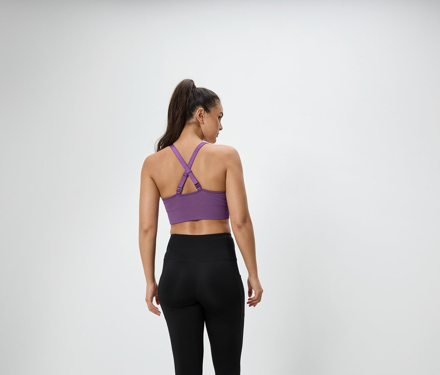 Egy barna hajú, lófarokba kötött nő háttal áll a kamerának, lila sportmelltartót és fekete leggingset visel.