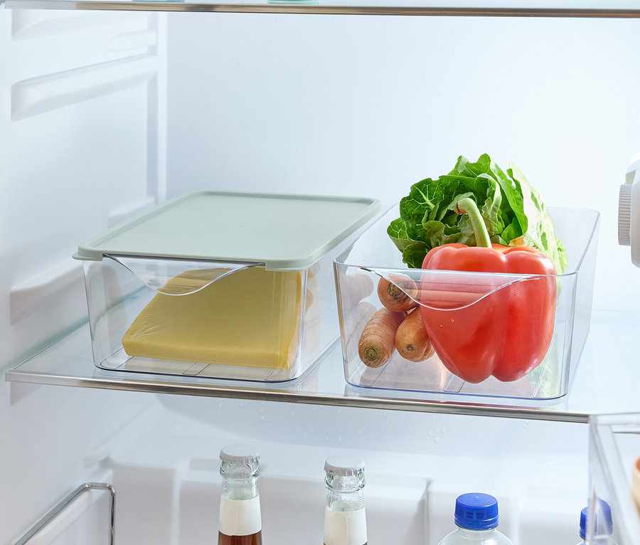 A hűtőszekrény polcain élelmiszerek találhatók: alma és citrom kerek tartókban, sajt műanyag dobozban fedővel, zöldségek (saláta, paprika, sárgarépa) műanyag dobozban és italos palackok egy tartóban.