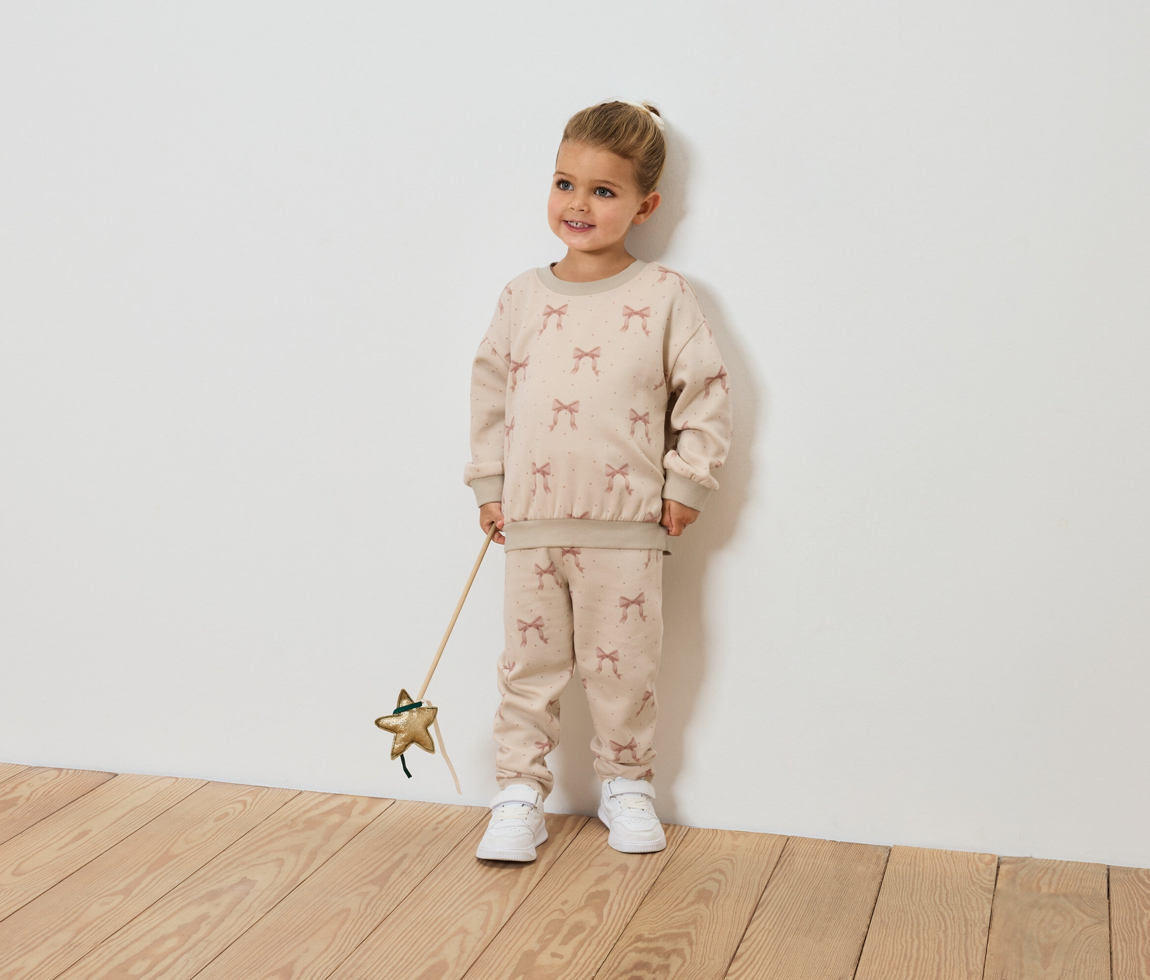 Egy mosolygó kislány csillag alakú varázspálcát tart, és bézs színű, masnis mintás ruhát visel.