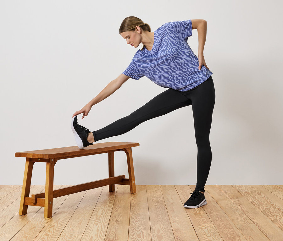 Kék pólót és fekete leggingset viselő nő nyújtózkodik, egyik lábát egy fa padra helyezve.