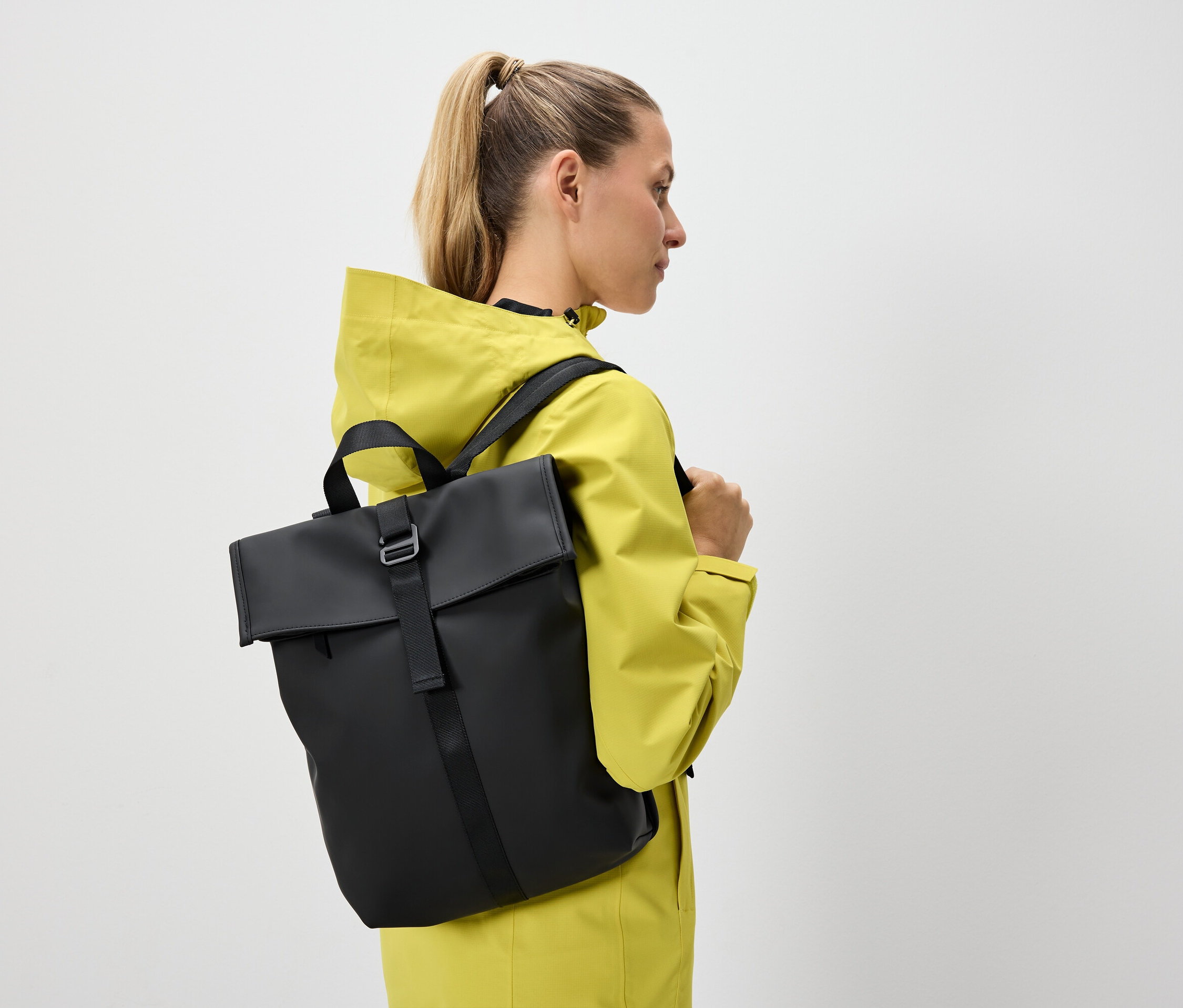 A szőke hajú, lófarokba kötött nő sárga kabátot és fekete hátizsákot visel.