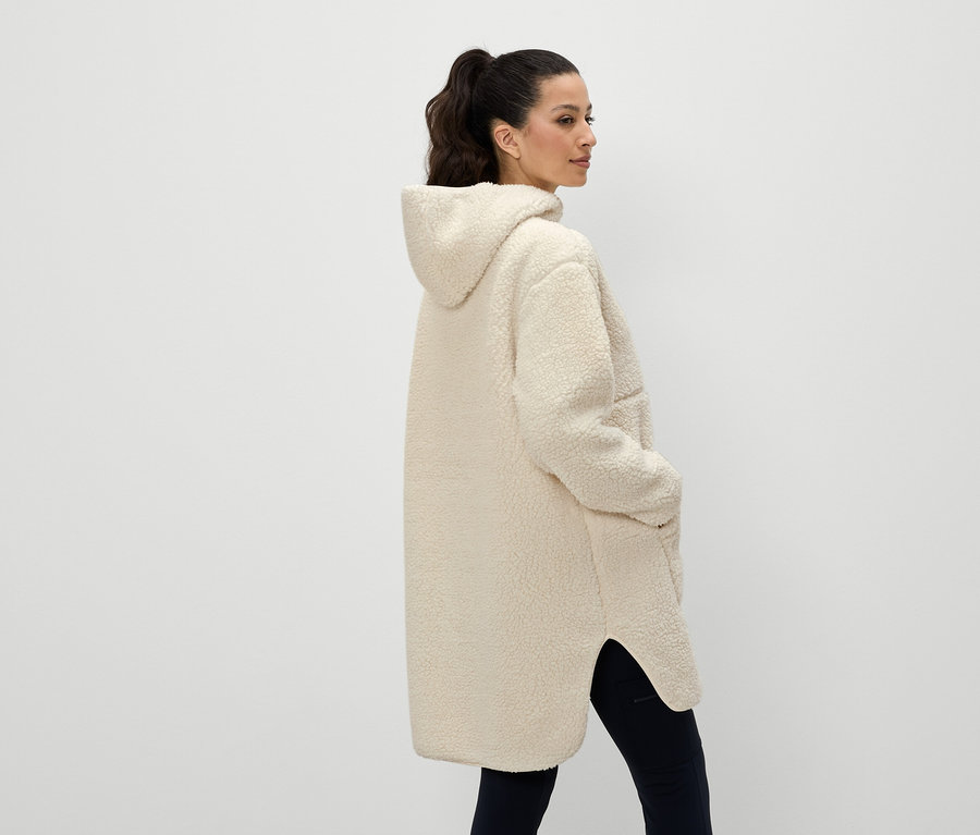 Hátulról egy nő krém színű kapucnis kabátot visel, keze a zsebében.
