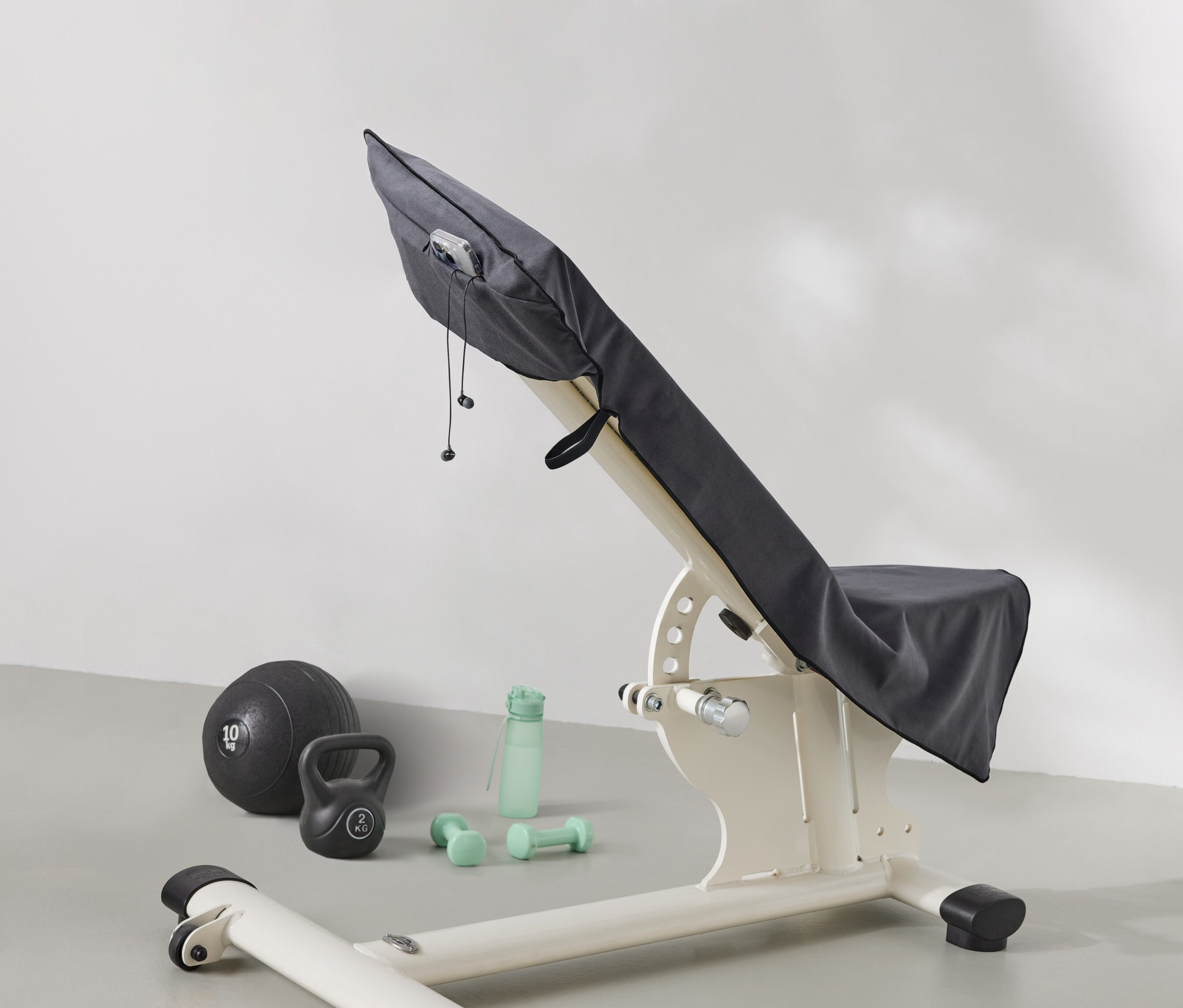 Egy súlyzópad szürke anyaggal van letakarva. A pad mellett egy medicinlabda, egy kettlebell, egy vizes palack és súlyzók találhatók.