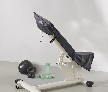 Egy súlyzópad szürke anyaggal van letakarva. A pad mellett egy medicinlabda, egy kettlebell, egy vizes palack és súlyzók találhatók.