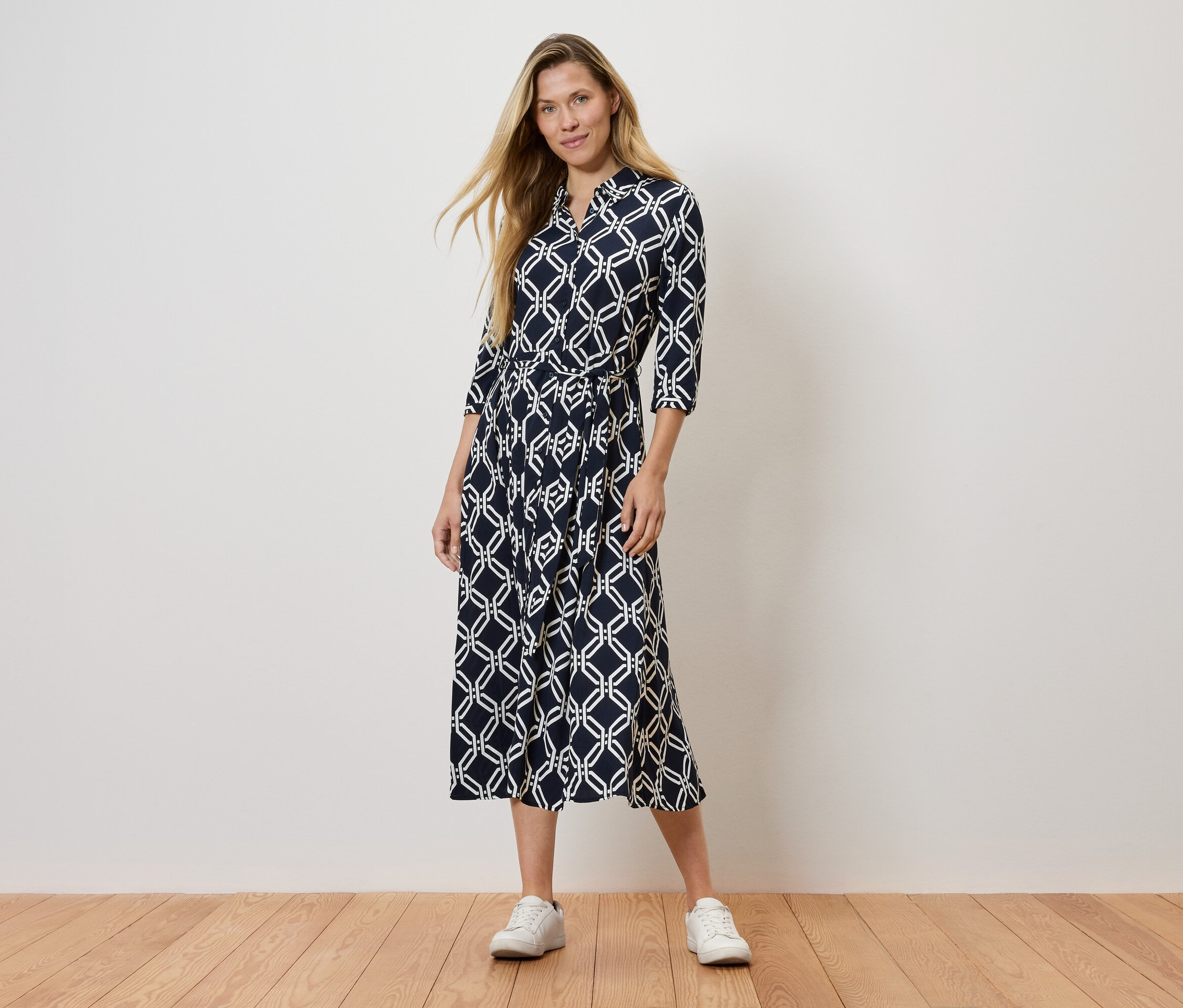 Egy nő áll egy sötétkék mintás ruhában és fehér tornacipőben.