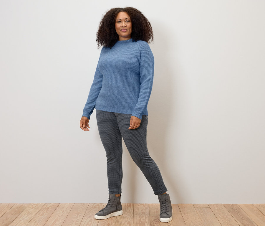 Fából készült padlón álló nő kék pulóvert és szürke, tyúkláb-jacquardmintás szövetnadrágot visel.