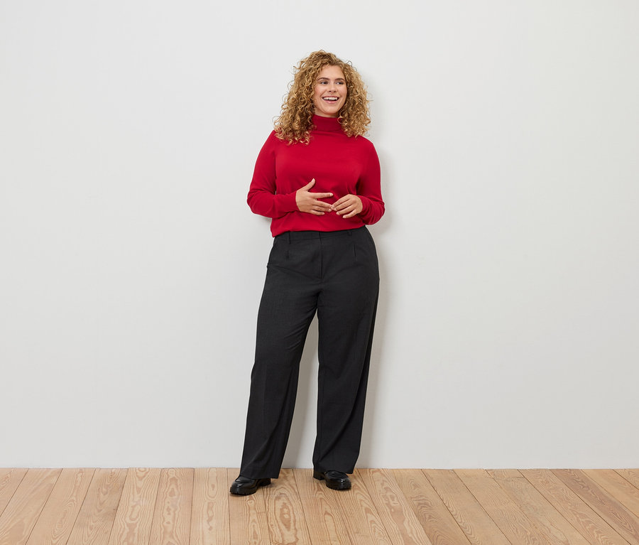 Egy göndör hajú nő áll, piros garbót és fekete nadrágot visel.