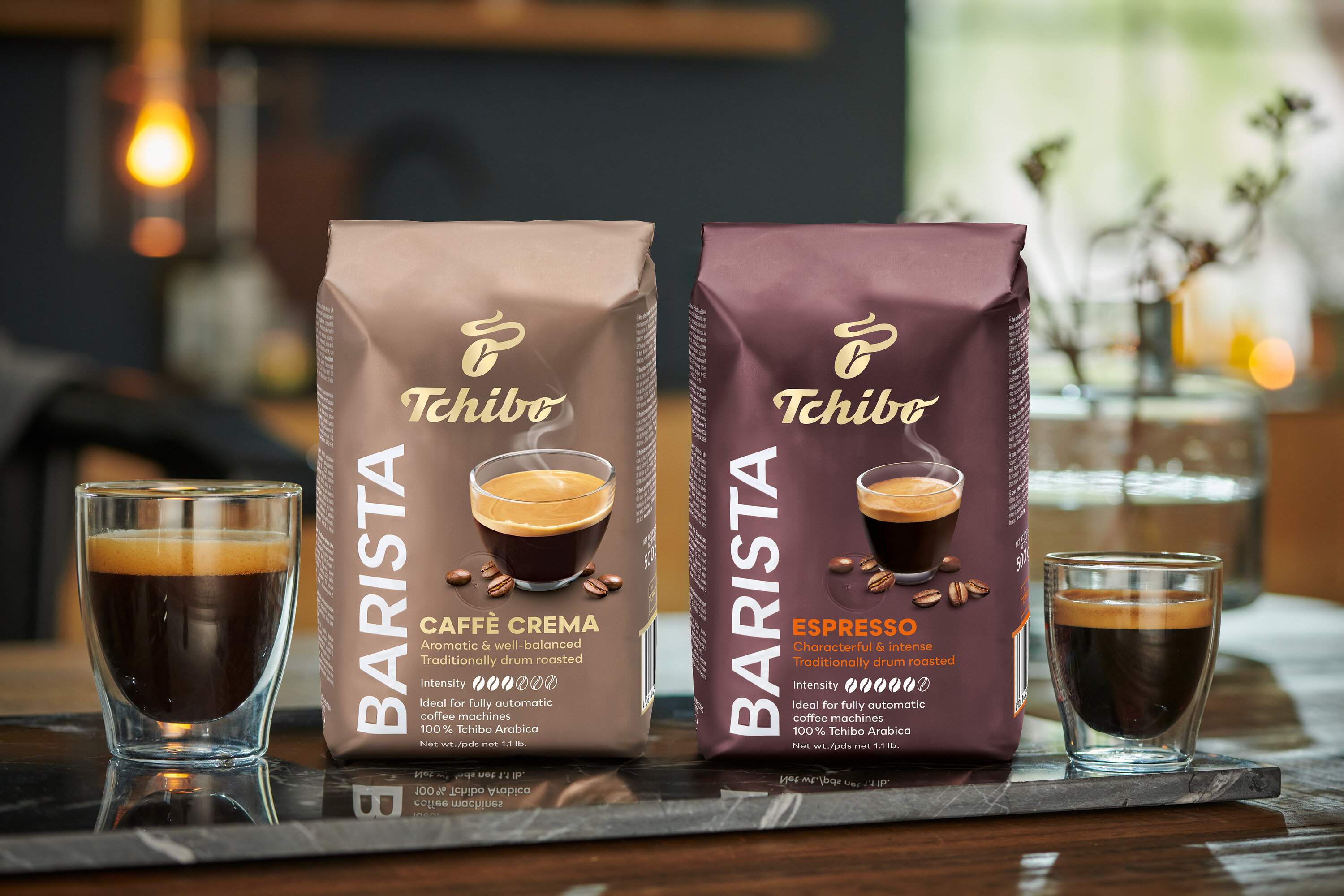 Két pohár kávé és két csomag Tchibo BARISTA Caffè Crema szemes kávé áll egy márványlapon.