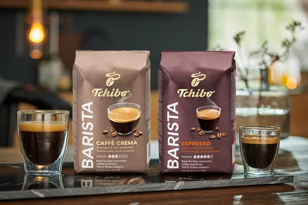 Két pohár kávé és két csomag Tchibo BARISTA Caffè Crema szemes kávé áll egy márványlapon.