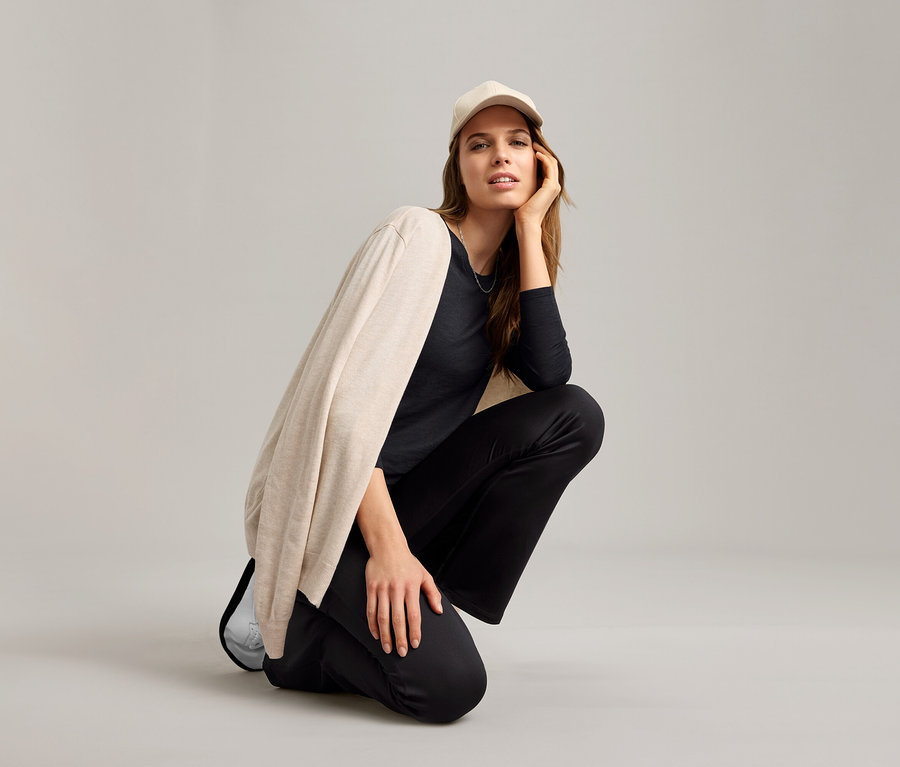 Nő guggol fekete funkcionális szabadidőfelsőben, bézs kardigánban és világos színű baseball sapkában.