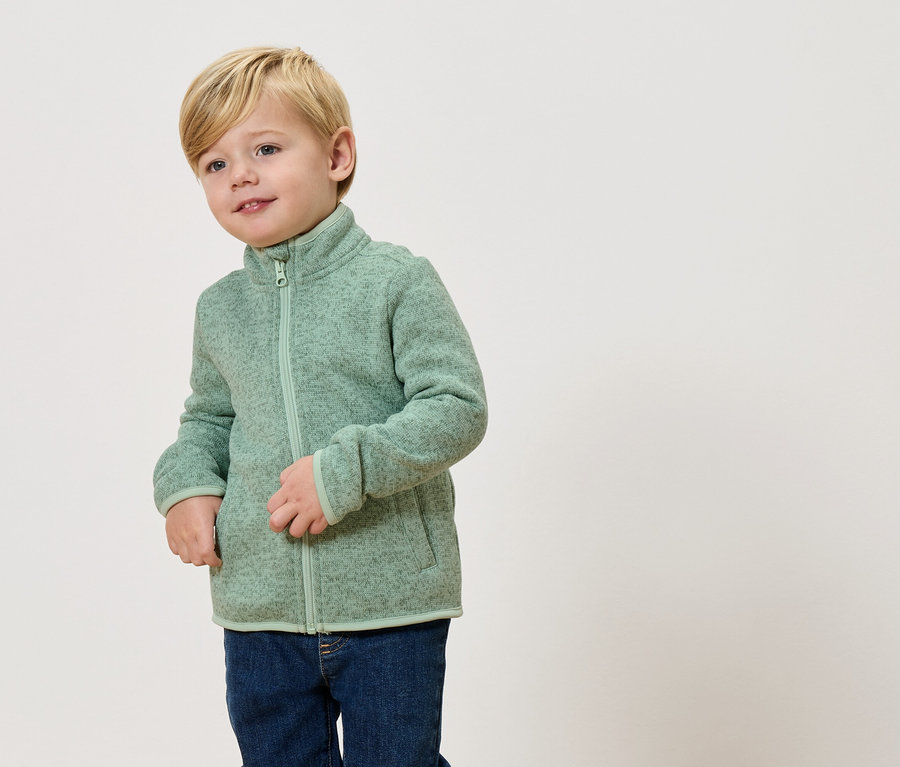 Egy szőke fiú világoszöld polár pulóvert és farmert visel.